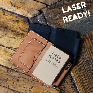 May include: A leather notebook cover in tan and black, with white stitching, holding a Field Notes memo book and a pen. The text "LASER READY!" is displayed in white. The notebook is on a wooden surface.