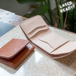May include: Two leather wallets, one open and one closed, sit on a stone surface. The open wallet is light tan, revealing card slots. The closed wallet is brown. Both have white stitching and the text "LASER READY!"