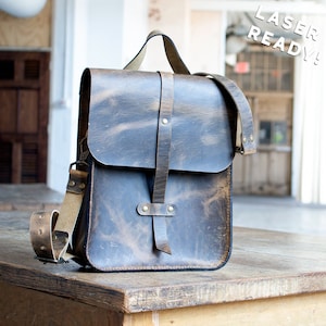 May include: A brown leather messenger bag with a flap closure and adjustable shoulder strap. The bag has a top handle and a vertical strap with rivets. The text "LASER READY!" is visible in the upper right corner.