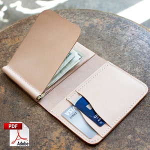 Könnte beinhalten: Offene, hellbraune Ledergeldbörse mit einem Geldscheinclip, der mehrere Geldscheine hält. Die Geldbörse hat Kartenfächer mit zwei sichtbaren Karten. Die Kanten sind mit hellem Faden genäht. Die Geldbörse ist geöffnet und zeigt ihren Inhalt.