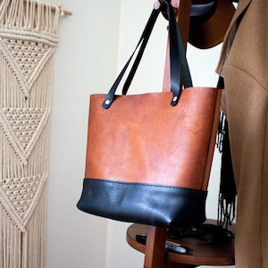 Puede incluir: Un bolso tote de cuero marrón y negro con asas negras cuelga de un perchero de madera. El bolso tiene forma rectangular con una sección superior marrón y una base negra. Las asas son negras y están sujetas con remaches plateados.