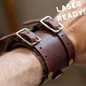 May include: A brown leather wrist cuff with two silver buckles and brass rivets. The cuff is wrapped around a wrist. The text "LASER READY!" is in white letters.