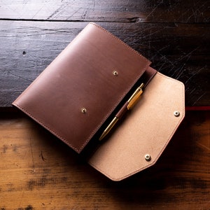 Könnte beinhalten: Ein braunes Leder-Portfolio mit einem goldenen Stift im Inneren. Das Portfolio ist geöffnet und zeigt ein hellbraunes Innenfutter und goldene Druckknöpfe. Der Stift ist goldfarben mit einem braunen Akzent.