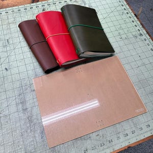 May include: Three leather journals in dark brown, bright red, and olive green, each secured with a colored elastic band. Beside them lies a rectangular, translucent acrylic template with small holes and the text "MAKE SUPPLY" etched in the corner.
