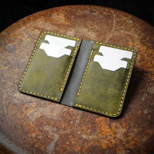 May include: Open leather card holder in olive green with yellow stitching. The wallet has two card slots, each holding a white card. The interior is gray. The wallet is open and displayed on a brown, textured surface.