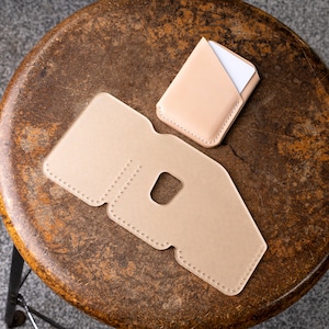 May include: Two pieces of light tan leather, one a card holder with a white card inside, the other a larger piece with a square cutout and dotted edges. The items are on a brown, distressed stool.