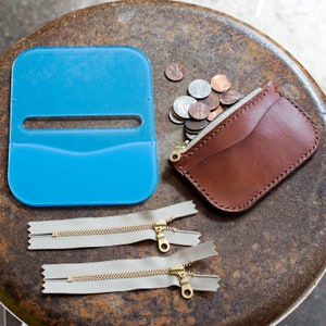 May include: A collection of craft supplies including a blue plastic template, two beige zippers with gold-colored metal teeth and pulls, a brown leather coin purse with a zipper, and a pile of US coins.
