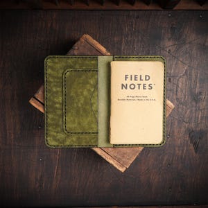 May include: An open, olive green leather notebook cover with black stitching, holding a "Field Notes" memo book. The memo book is tan with black text. The cover is on a wooden surface.