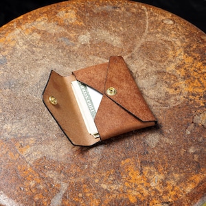May include: A brown leather wallet, open to reveal folded bills. The wallet has a triangular design with a snap closure. The leather has a textured appearance, and the wallet is set on a brown surface.