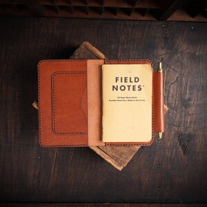 May include: A brown leather notebook cover with a matching pen holder, holding a "Field Notes" memo book. The memo book is cream-colored with black text. The leather has visible stitching and is set on a wooden surface.