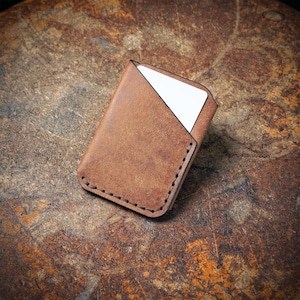 May include: A brown leather card holder with a stitched border. The holder is open, revealing a white card inside. The leather has a natural, textured appearance. The card holder is on a textured, brown surface.