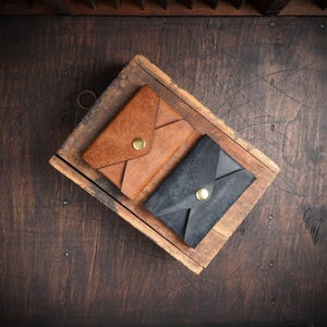 May include: Two leather envelope wallets, one brown and one black, are displayed on a wooden tray. Each wallet has a flap closure secured by a gold-colored snap. The wallets are set against a dark, textured wooden surface.