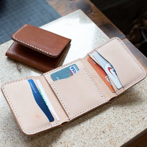 May include: Two leather wallets. One is a bi-fold in a light brown color, and the other is a tri-fold wallet in a natural tan color. The tri-fold wallet is open, displaying several credit cards in its pockets. White stitching is visible.