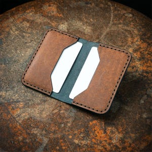 May include: Open brown leather wallet with two white card slots. The wallet has a dark brown border with black stitching. The interior is a dark gray color. The wallet is open and displayed on a textured brown surface.