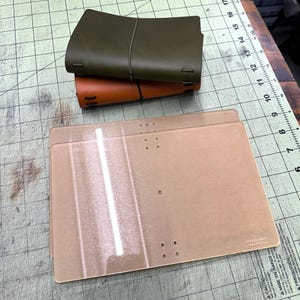 May include: An overhead shot shows two leather journals, one olive green and one brown, stacked on top of each other. Below them are two clear acrylic templates with pre-drilled holes, likely for crafting. The items are on a green cutting mat.