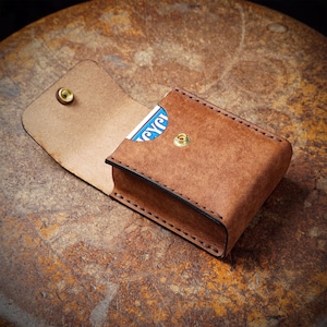 May include: A brown leather card case with a snap closure, open to reveal a playing card. The case has a stitched border and a brass-colored snap. The leather has a textured appearance.