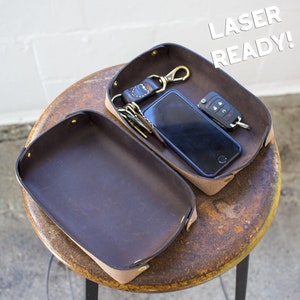 Può includere: Due vassoietti in pelle marrone con rivetti in ottone. Un vassoio contiene uno smartphone, chiavi e un portachiavi per auto. L'altro vassoio è vuoto. Il testo "LASER READY!" è sullo sfondo.