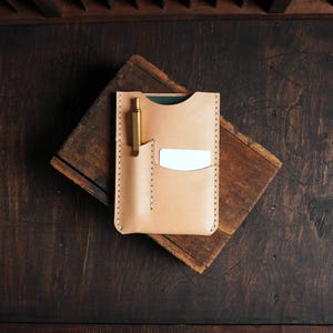 May include: A light tan leather cardholder with a pen and a white card in the pockets. The cardholder is stitched with a contrasting thread and sits on a dark wooden surface. A gold-colored pen is in the pen pocket.
