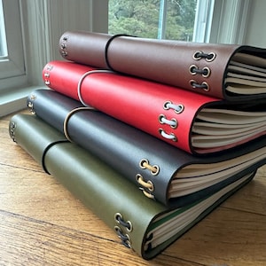 May include: A stack of leather-bound journals in various colors: brown, red, black, and olive green. Each journal has a wrap-around closure and decorative eyelets along the spine. The pages are visible, suggesting a notebook or diary.