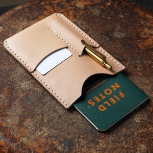 May include: A light tan leather wallet with a card slot, a pen holder, and a small green notebook with the words "FIELD NOTES" in orange. A gold-colored pen is in the pen holder. The wallet is on a brown surface.