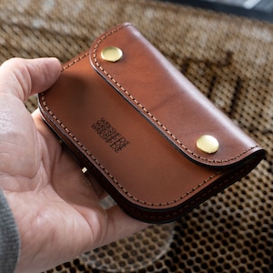 May include: A brown leather wallet with a curved flap secured by two brass snap closures. The wallet has stitched edges and the text "WRKSHP.EYE" is printed on the front. The wallet is held in a hand.