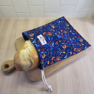 May include: A rectangular fabric bread bag with a dark blue background featuring a colorful floral pattern in red, yellow, and light blue. The bag has a white drawstring closure and a white label with the text Aunt Me Crafts. A loaf of bread is inside.