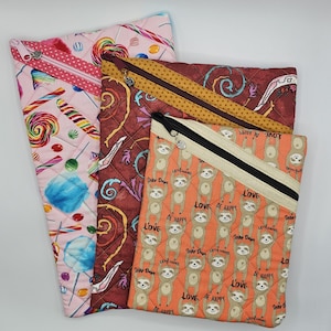 May include: Three quilted zippered pouches with different patterns. The first pouch is pink with a candy cane and lollipop pattern. The second pouch is brown with a swirling pattern. The third pouch is orange with a sloth pattern and the text "Love, Be Happy, Let's Hang, Toby Days".