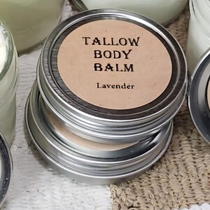 Può includere: Una pila di piccoli barattoli d'argento con etichette marroni. Le etichette riportano "TALLOW BODY BALM" e "Lavender" in testo nero. I barattoli sono impilati su una superficie strutturata dai toni neutri, suggerendo un prodotto naturale per la cura della pelle.