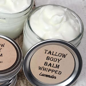 Op de afbeelding: Close-up van verschillende kleine glazen potjes gevuld met witte, opgeklopte body balm. De zilveren deksels hebben een bruin label met de tekst "Tallow Body Balm Whipped Lavender". De potjes staan op een getextureerd oppervlak.