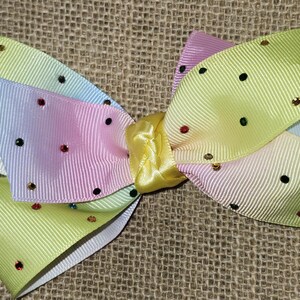 Può includere: Un grande fiocco per capelli multistrato con un centro giallo e nastro arcobaleno pastello. Il fiocco è decorato con piccoli strass colorati. Il fiocco misura circa 15 cm di larghezza.