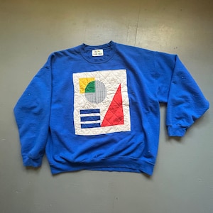 May include: A royal blue sweatshirt with a geometric design. The design features a white square with a yellow square, a green semi-circle, a black and white striped circle, a red triangle, and blue horizontal lines. The sweatshirt has a crew neck.