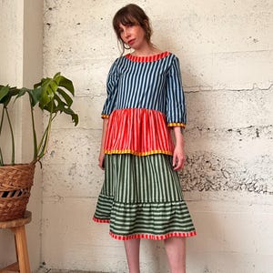 以下が含まれることがあります： 丸いネックラインと七分袖のカラフルなストライプ柄のワンピース。 上部は青と白のストライプで、赤いミドルセクション、緑と白のストライプのスカートに変わります。 ワンピースはティアードデザインで、黒い靴を履いています。