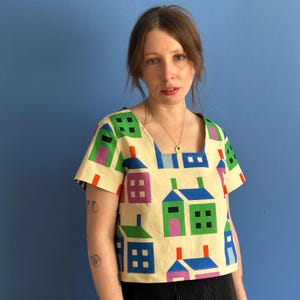 May include: A cream-coloured, square-neck top featuring a repeating pattern of colourful houses. The houses are in shades of green, blue, purple, and pink, with orange and red accents. The top has short sleeves.