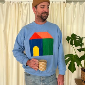 May include: Light blue sweatshirt featuring a colorful house design with a red roof, blue walls, a green side, and a yellow door. The person is wearing a brown beanie and holding a checkered mug. The background includes a plant and cream-colored curtains.
