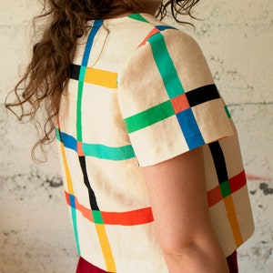 May include: Cream-colored short-sleeved top with a colorful grid pattern. The grid features vertical and horizontal lines in blue, green, red, yellow, and black. The top is paired with a red bottom, creating a striking contrast. The top is made of a lightweight fabric.