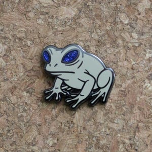 May include: A light gray frog enamel pin with black outlines. The frog has large eyes filled with blue glitter. The pin is set against a textured, brown background. The frog is a whimsical accessory.