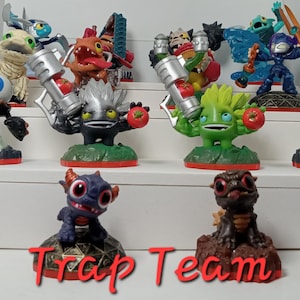May include: A collection of 11 colorful Skylanders Trap Team figures, including a green and red figure with a large eye, a black and white figure with a large eye, a black and red figure with a large eye, a black and white figure with a large eye, a black and white figure with a large eye, a black and white figure with a large eye, a black and white figure with a large eye, a black and white figure with a large eye, a black and white figure with a large eye, a black and white figure with a large eye, and a black and white figure with a large eye. The figures are arranged on three shelves, with the text "Trap Team" printed in red letters below the figures.