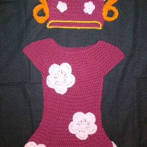 May include: A crocheted dress and hat set in pink and burgundy with white flower accents. The hat has two orange yarn pigtails.