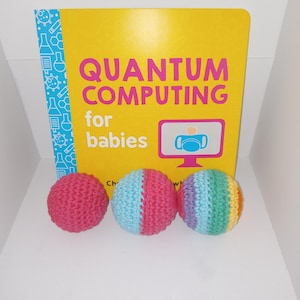 May include: Three crocheted balls, one pink, one blue and pink, and one rainbow colored. The balls are in front of a yellow book with the title "Quantum Computing for babies".