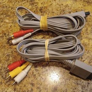 May include: Two gray AV cables, each coiled and secured with a rubber band. One cable has red, white, and yellow connectors, the other has red, white, and yellow connectors. The cables are for a gaming console.