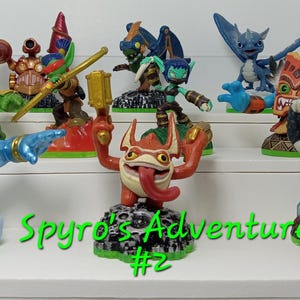 May include: A collection of colorful toy figures, including a blue and red figure with a long tongue, a green figure with a red rocket, a blue figure with a shark fin, and a red and orange figure with a tiki mask. The figures are arranged on a white shelf with a green background. The text "Spyro's Adventure #2" is written in green letters below the figures.