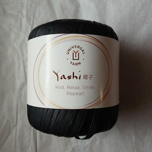 May include: A black yarn ball with a white label wrapped around it. The label features the text "Yashi" in a stylized font, along with the words "Knit, Relax, Smile, Repeat!" and the Universal Yarn logo.
