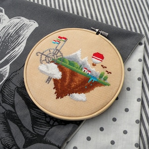 Peut inclure: Un cerceau de broderie beige avec un motif de point de croix représentant une île flottante avec des montagnes, une maison, des arbres, une montgolfière et des nuages. L'île est brune avec de l'herbe verte et une rivière bleue.