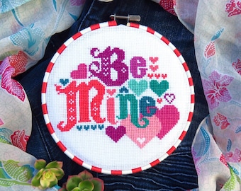 Peut inclure: Un cercle de broderie au point de croix avec les mots "Be Mine" en rouge, rose et violet, entouré de cœurs. Le cercle a une bordure rayée rouge et blanche. Le fond comprend du denim et une écharpe à motifs floraux.