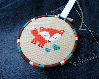 May include: A brown embroidery hoop with a white fabric background featuring a red fox with closed eyes and two teal hearts. The hoop is decorated with a teal, orange, and white striped border.