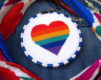 May include: A white embroidery hoop with a rainbow heart design on a blue denim background. A colorful scarf with red, white, and blue accents is draped over the denim.