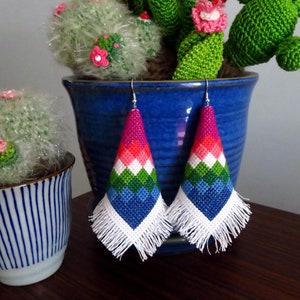 May include: A pair of colorful, triangular earrings with white fringe. The earrings are made of cross-stitch fabric and feature a geometric pattern of pink, red, white, green, and blue.