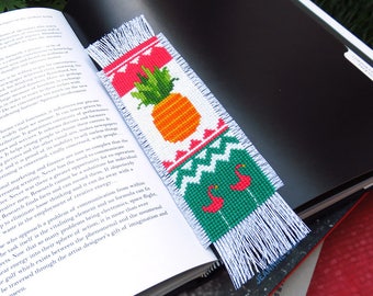 May include: A handmade bookmark with a cross-stitch design featuring a pineapple, flamingos, and a geometric pattern. The bookmark has a white fringe and is placed on an open book.