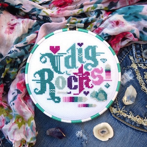 May include: A white embroidery hoop with a green and white border. The embroidery reads "Dig Rocks!" in a stylized font with pink, purple, and green accents.