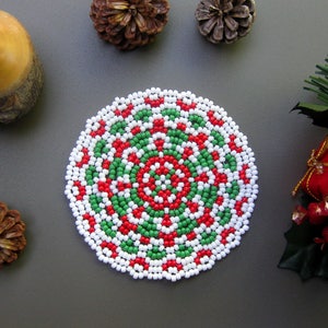 May include: A round, white, green, and red beaded coaster with a circular pattern. The coaster is made of small beads that are sewn together to create a decorative design.